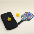 A pickleball set with two yellow hybrid pickleballs, one placed in the outer net compartment of a black bag, a paddle with modern, artistic patterns in bold, vibrant colors lying flat, and the black carry bag beside it, showcasing the unique style of the gear.