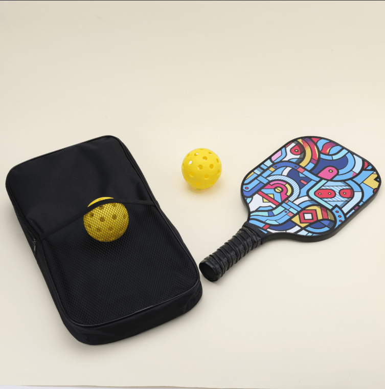 A pickleball set with two yellow hybrid pickleballs, one placed in the outer net compartment of a black bag, a paddle with modern, artistic patterns in bold, vibrant colors lying flat, and the black carry bag beside it, showcasing the unique style of the gear.