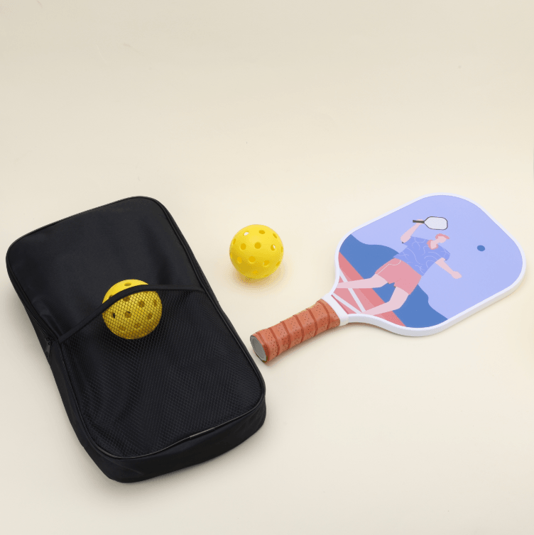 A pickleball set with two yellow hybrid pickleballs, one placed in the outer net compartment of a black bag, a paddle with vibrant, fun, sports-themed patterns and bold colors lying flat, and the bag beside it.